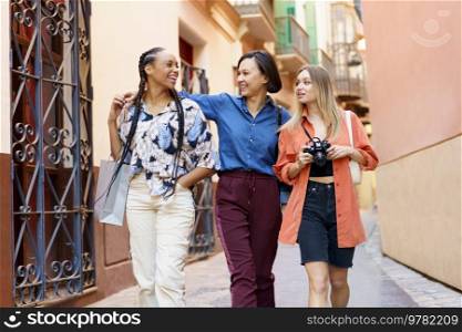Blond woman with photo camera in hands walking near urban building with diverse friends while looking at each other delightfully. Happy multiracial ladies with camera against building