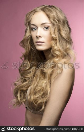 Blonde beautiful young girl with long curly hair in beige lingerie on pink backgroung is looking in camera