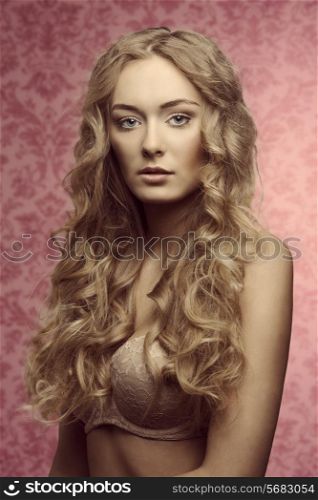 Blonde beautiful young girl with long curly hair in beige lingerie on on pink vintage background looking in camera