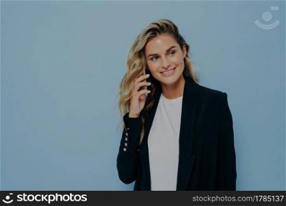 Blonde business woman in black jacket talking on mobile phone with family, happy to hear good news, listening and looking sideways while standing isolated on blue background. Communication concept. Blonde woman talking on phone with her relatives