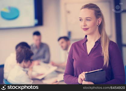 Blonde businesswoman in casual clothes working on tablet computer at modern startup business office interior. Young people group on team meeting blured in background