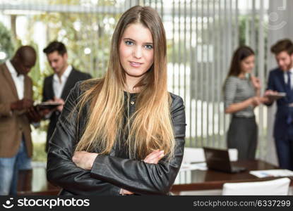 Blonde businesswoman leader looking at camera with arms crossed in working environment. Group of people in the background