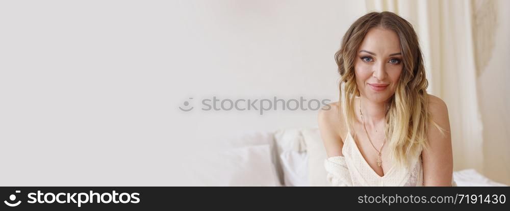 Blonde European female sitting on a bed in a bright modern bedroom. Models skin without retouching, the concept of naturalness. Banner with copy space, selective focus