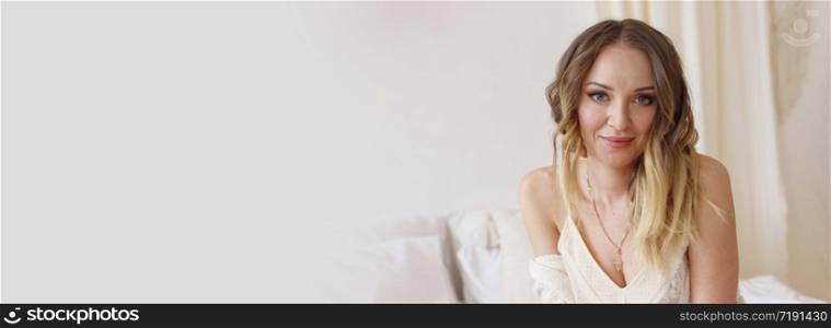 Blonde European female sitting on a bed in a bright modern bedroom. Models skin without retouching, the concept of naturalness. Banner with copy space, selective focus