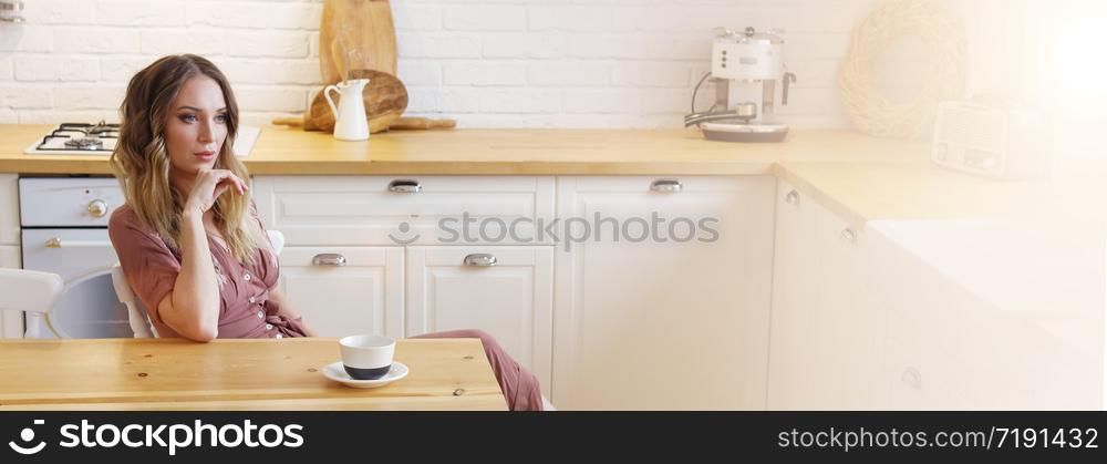 Blonde European female wearing beige dress sitting in bright modern kitchen. Banner with selective focus. Quarantine. Self isolation. Stay home concept.