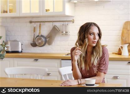 Blonde European female wearing beige dress sitting in bright modern kitchen holding cup in her hands. Picture with selective focus. Quarantine. Self isolation. Stay home concept.