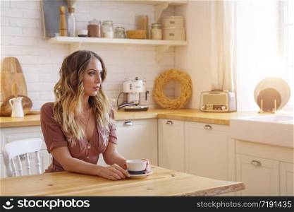 Blonde European female wearing beige dress sitting in bright modern kitchen. Picture with selective focus. Quarantine. Self isolation. Stay home concept.