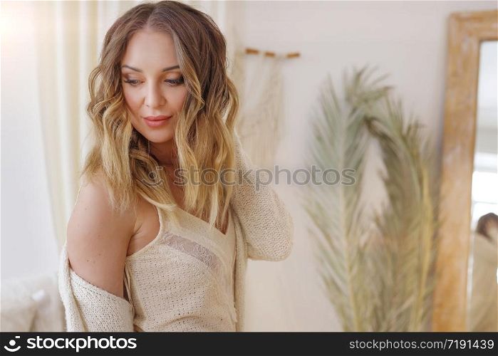 Blonde European female wearing tender fashionable cardigan sitting in bright modern bedroom with big mirror. Picture with selective focus. quarantine, stay home