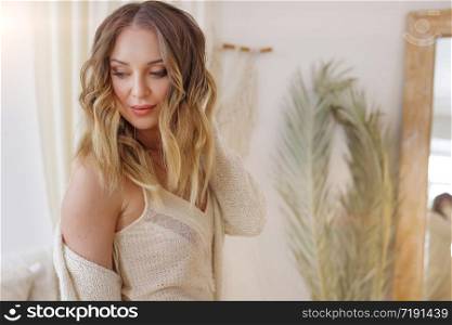Blonde European female wearing tender fashionable cardigan sitting in bright modern bedroom with big mirror. Picture with selective focus. quarantine, stay home