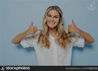 Blonde female dressed in white shirt expressing positive emotions while showing thumbs up gesture with both of her hands, displaying that she likes it, isolated next to blue wall. Blonde girl showing thumbs up gesture with both hands