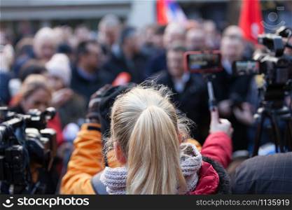 Blonde female reporter covering media event, filming press or news conference with smartphone