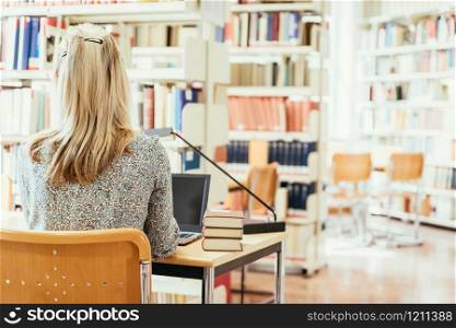 Blonde female student is sitting at the desk with pile of books, university library