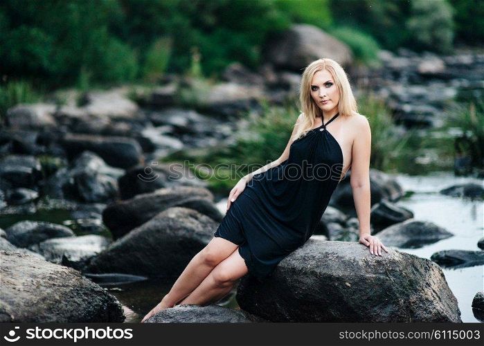 blonde girl in a black dress with blue eyes on the stone coast of the river