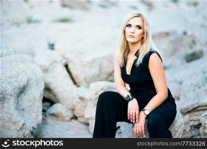 blonde girl in a black trouser suit with blue eyes on the background of a granite quarry from gray rubble