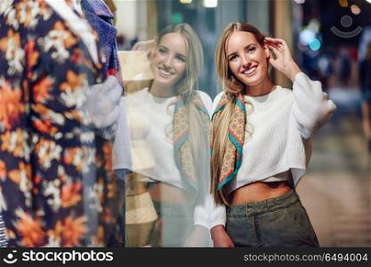 Blonde girl smiling with defocused urban city lights at night. Blonde girl wearing white sweater smiling in the street with defocused city lights at the background. Pretty woman with pigtail hairstyle at night reflected in a shop window.