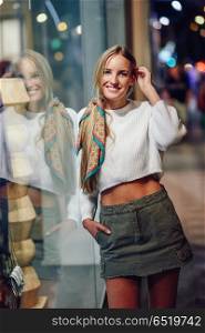Blonde girl smiling with defocused urban city lights at night. Blonde girl wearing white sweater smiling in the street with defocused city lights at the background. Pretty woman with pigtail hairstyle at night reflected in a shop window.