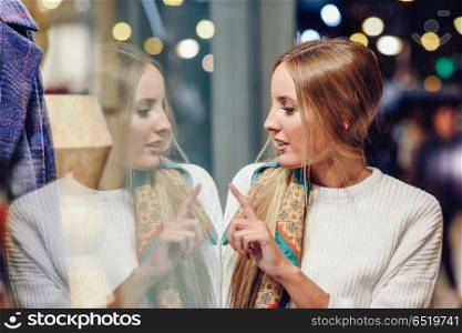 Blonde girl with defocused urban city lights at night. Blonde girl wearing white sweater standing in the street with defocused city lights at the background. Pretty woman with pigtail hairstyle at night reflected in a shop window.