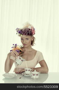 blonde girl with floral decoration and classical tea set on the table, looks in to the lens and takes the milk jug with right hand