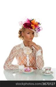 blonde girl with floral hat and vintage shirt behind the table with cake and cup of tea, her face is turned at left and has the left hand under the chin