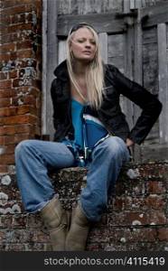 Blonde in denim sits on brick wall outside disused warehouse