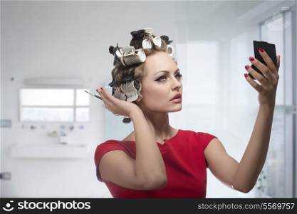 Blonde lady taking care of her style and checking that in the little mirror. Applying make-up with brush and styling her hair with curlers, wearing fashion red dress