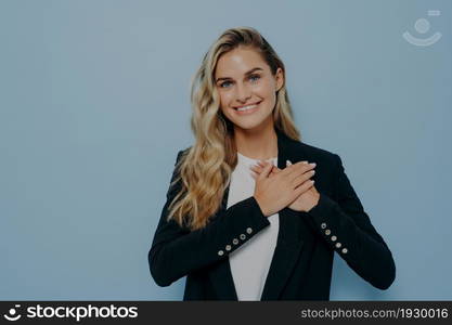 Blonde positive young female in black coat showing her appreciation and gratitude by placing hands on chest, thanking with gesture, standing isolated on blue background, copy space for text. Cheerful blonde girl showing her appreciation and gratitude by placing hands on chest