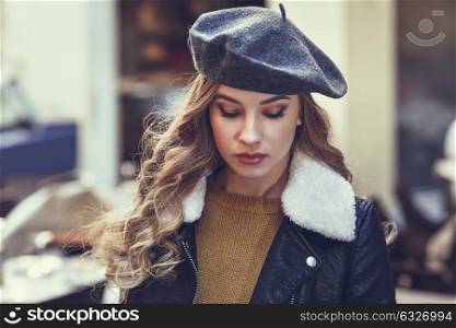 Blonde russian woman in urban background. Beautiful young girl wearing beret, black leather jacket and mini skirt standing in the street. Pretty female with long wavy hair hairstyle.