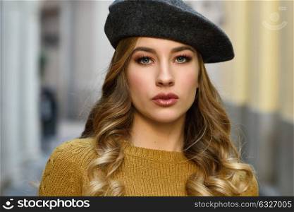 Blonde russian woman in urban background. Beautiful young girl wearing beret, black leather jacket and mini skirt standing in the street. Pretty female with long wavy hair hairstyle and blue eyes.
