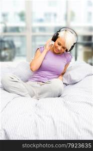 Blonde smiling woman is listening to music with headphones in the bed