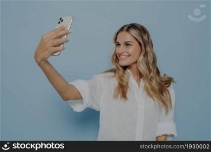 Blonde teenage girl taking selfie on modern smartphone for social media account with bright smile, smiling for camera and sharing photos with her friends, standing isolated next to blue background. Blonde teenage girl taking selfie while smiling