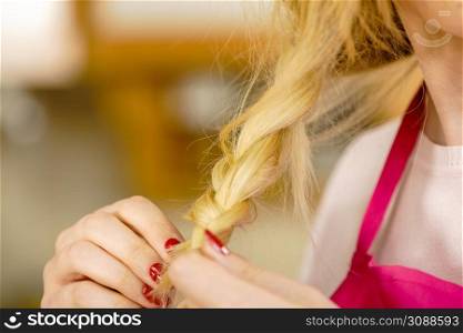 Blonde woman creating her hairstyle, making braid wearing apron. Lovely female getting ready. Waitress, chef getting ready to work. Woman making braid on blonde hair