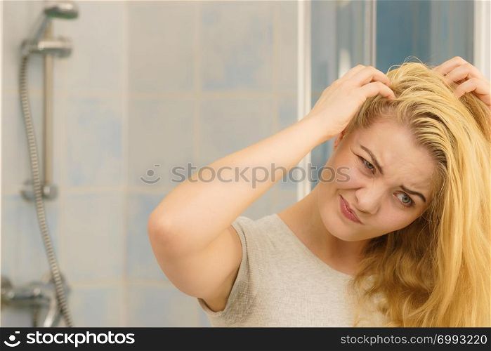 Blonde woman having problems with greasy oily hair looking at herself in bathroom. Female showing scalp, scratching herself, dandruff problem.. Female having problem with blonde hair