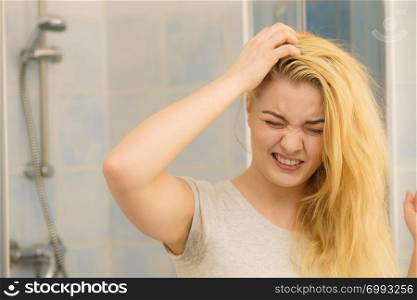Blonde woman having problems with greasy oily hair looking at herself in bathroom. Female showing scalp, scratching herself, dandruff problem.. Female having problem with blonde hair