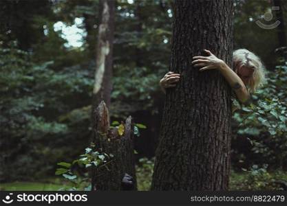 Blonde woman hiding behind old tree scenic photography. Picture of person with wild forest on background. High quality wallpaper. Photo concept for ads, travel blog, magazine, article. Blonde woman hiding behind old tree scenic photography