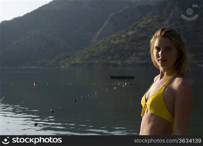 Blonde woman in bikini looks back at camera in morning sunlight of lake