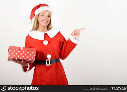 Blonde woman in Santa Claus clothes smiling with gift box in her hands. Young female with blue eyes pointing right with her finger, isolated on white