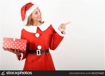 Blonde woman in Santa Claus clothes smiling with gift box in her hands. Young female with blue eyes pointing right with her finger, isolated on white