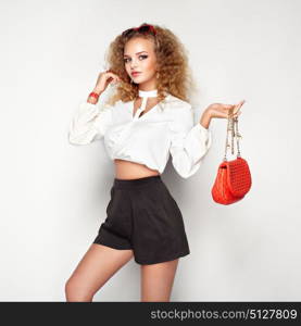 Blonde woman in summer blouse and shorts. Girl posing on white background. Red handbag. Stylish curly hairstyle. Glamour lady in stylish sunglasses