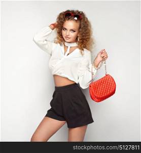 Blonde woman in summer blouse and shorts. Girl posing on white background. Red handbag. Stylish curly hairstyle. Glamour lady in stylish sunglasses