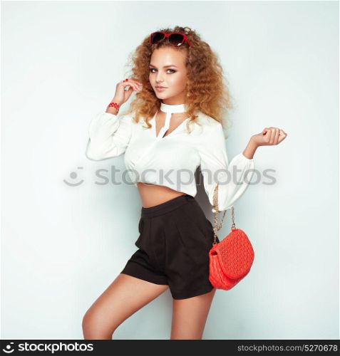 Blonde woman in summer blouse and shorts. Girl posing on white background. Red handbag. Stylish curly hairstyle. Glamour lady in stylish sunglasses