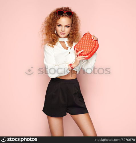 Blonde woman in summer blouse and shorts. Girl posing on white background. Red handbag. Stylish curly hairstyle. Glamour lady in stylish sunglasses