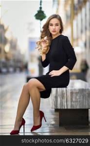 Blonde woman in urban background. Beautiful young girl wearing black elegant dress and red high heels standing in the street. Pretty russian female with long wavy hair hairstyle and blue eyes.