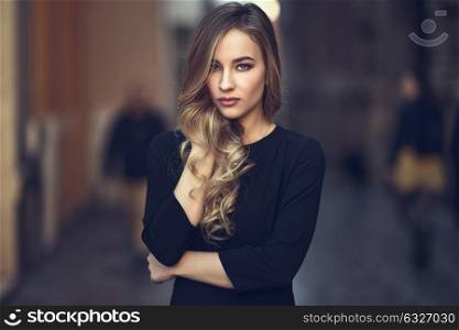 Blonde woman in urban background. Beautiful young girl wearing black elegant dress standing in the street. Pretty russian female with long wavy hair hairstyle and blue eyes.