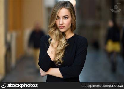 Blonde woman in urban background. Beautiful young girl wearing black elegant dress standing in the street. Pretty russian female with long wavy hair hairstyle and blue eyes.