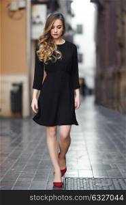 Blonde woman in urban background. Beautiful young girl wearing black elegant dress and red high heels standing in the street. Pretty russian female with long wavy hair hairstyle.