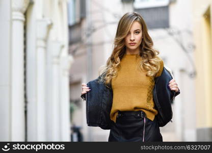 Blonde woman in urban background. Beautiful young girl wearing black leather jacket and mini skirt standing in the street. Pretty russian female with long wavy hair hairstyle and blue eyes.