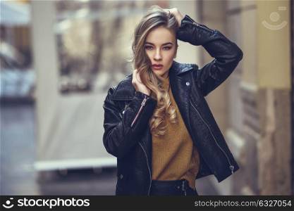 Blonde woman in urban background. Beautiful young girl wearing black leather jacket and mini skirt standing in the street. Pretty russian female with long wavy hair hairstyle and blue eyes.
