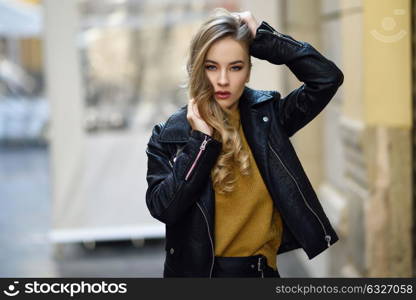 Blonde woman in urban background. Beautiful young girl wearing black leather jacket and mini skirt standing in the street. Pretty russian female with long wavy hair hairstyle and blue eyes.