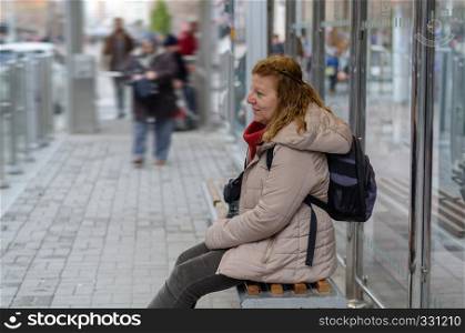 Blonde woman is waiting for the tram at the stop.She is tired and is sitting on the bench.