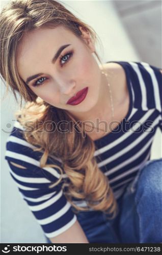 Blonde woman, model of fashion, possing in urban background.. Close-up portrait of blonde woman, model of fashion, possing in urban background. Beautiful young girl wearing striped t-shirt and blue jeans in the street. Pretty russian female with pigtail.